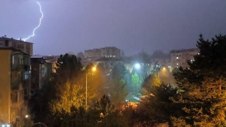 Gece karanlığında apartmanların üzerinde şimşek çakarken, ağaçlarla çevrili sokaklar ve yanan lambalar görülüyor.