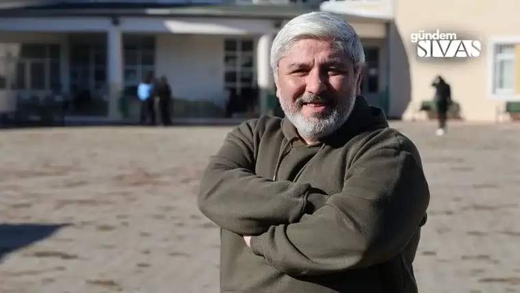 Kahverengi bir kapüşonlu giysi giymiş, gülümseyen bir adam, kollarını kavuşturmuş.