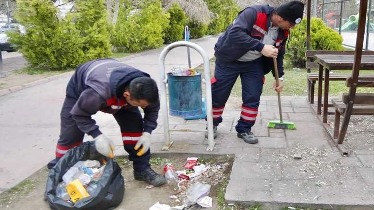 İki temizlik görevlisi, parkta yere saçılmış çöpleri eldiven ve süpürgeyle topluyor.