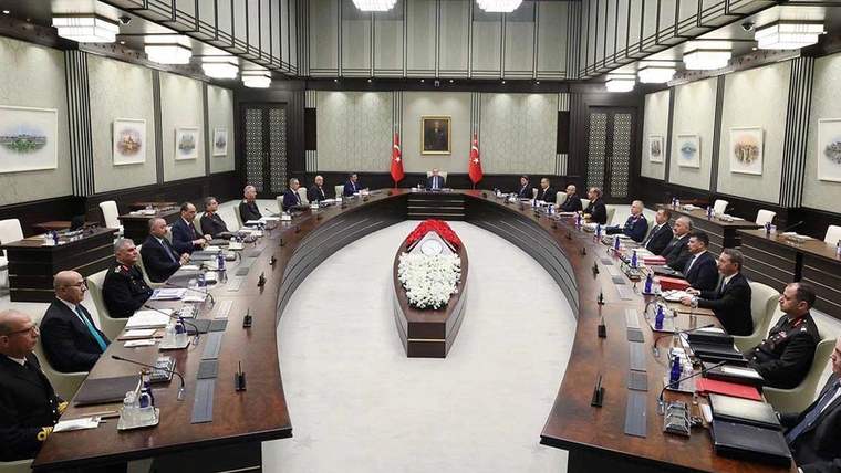 Toplantı masasında oturan birçok kişi, masanın ortasında beyaz çiçekler yer alıyor.