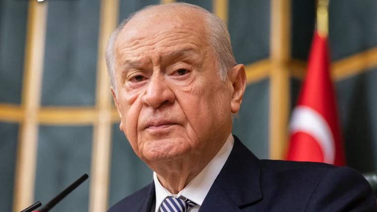 Takım elbiseli yaşlı bir erkek, önündeki mikrofonların yanında duruyor; arka planda Türk bayrağı görülüyor.
