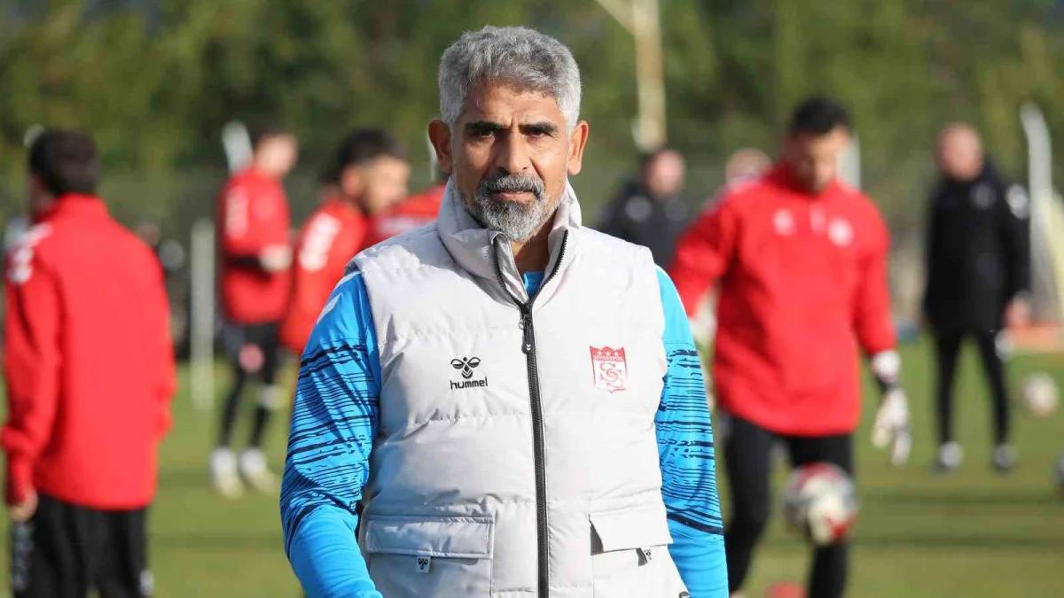 Gri saçlı bir kişi, beyaz yelek ve mavi üstle futbol sahasında duruyor; arka planda kırmızı formalı kişiler var.