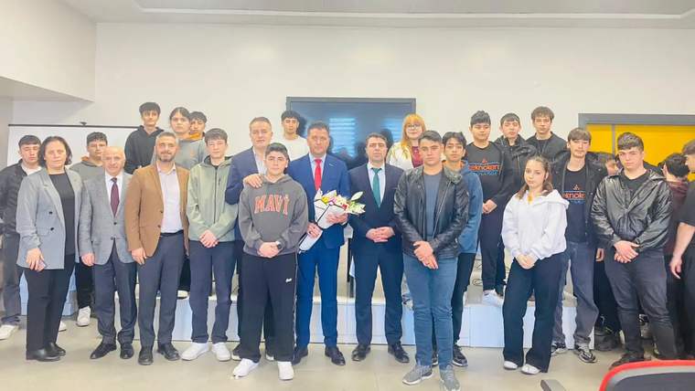 Bir grup öğrenci ve yetişkin, bir etkinlikte poz veriyor; ön planda çiçek tutan bir kişi var.
