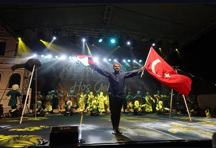 Sahnede iki bayrak tutan bir kişi, arkasında geleneksel kıyafetli dansçıların bulunduğu ışıklı alanda duruyor.