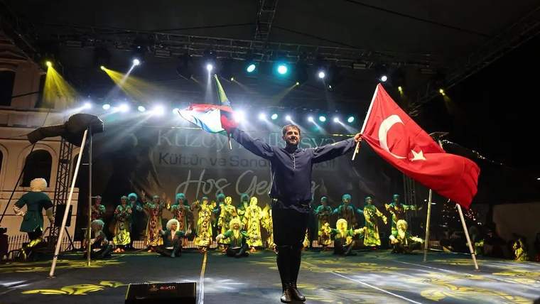 Sahnede iki bayrak tutan bir kişi, arkasında geleneksel kıyafetli dansçıların bulunduğu ışıklı alanda duruyor.