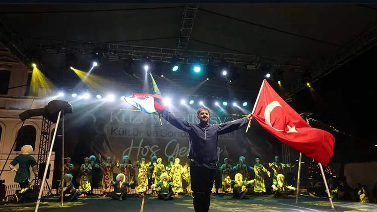 Sahnede iki bayrak tutan bir kişi, arkasında geleneksel kıyafetli dansçıların bulunduğu ışıklı alanda duruyor.