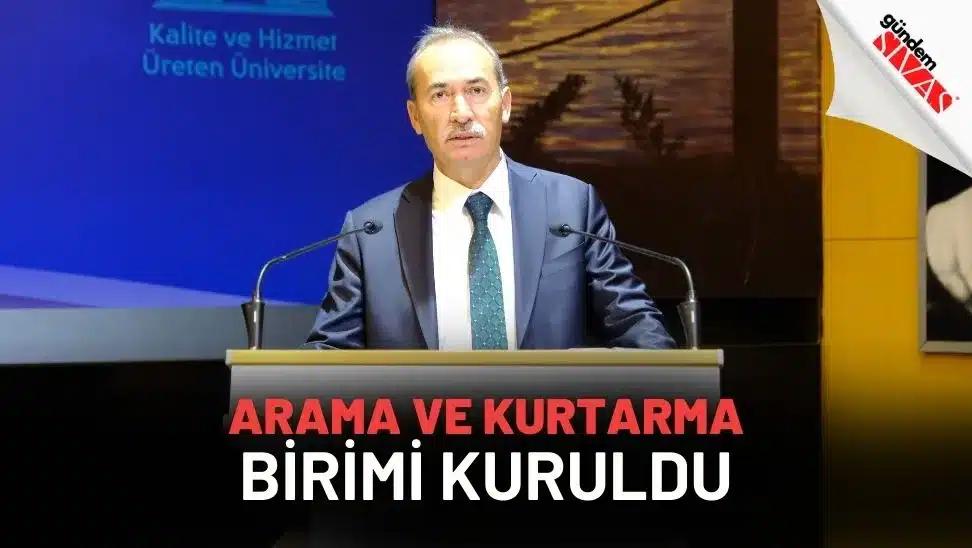Kürsüde konuşan takım elbiseli bir kişi, iki mikrofon arasında; arka planda üniversite sloganı görülüyor.