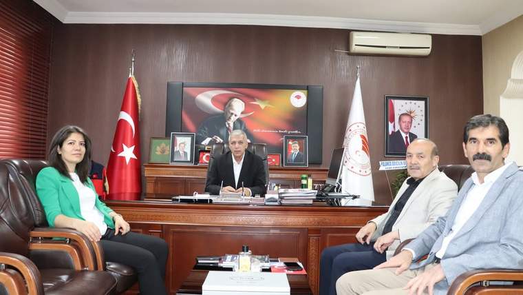 Masa arkasında oturan bir kişi ile karşısındaki koltuklarda oturan dört kişi, Türk bayrağı ve kurum flamalarıyla birlikte görülüyor.