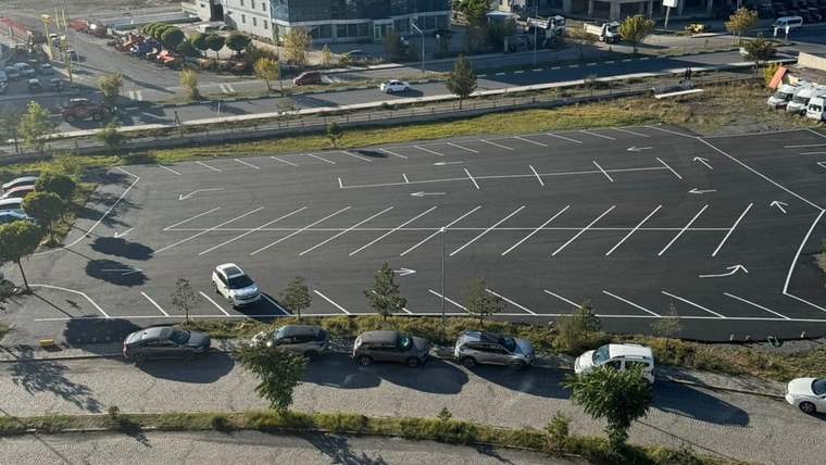 Çizgileri yenilenmiş geniş otoparkta birkaç otomobil, çevresindeki yollarda ise araçlar görülüyor.