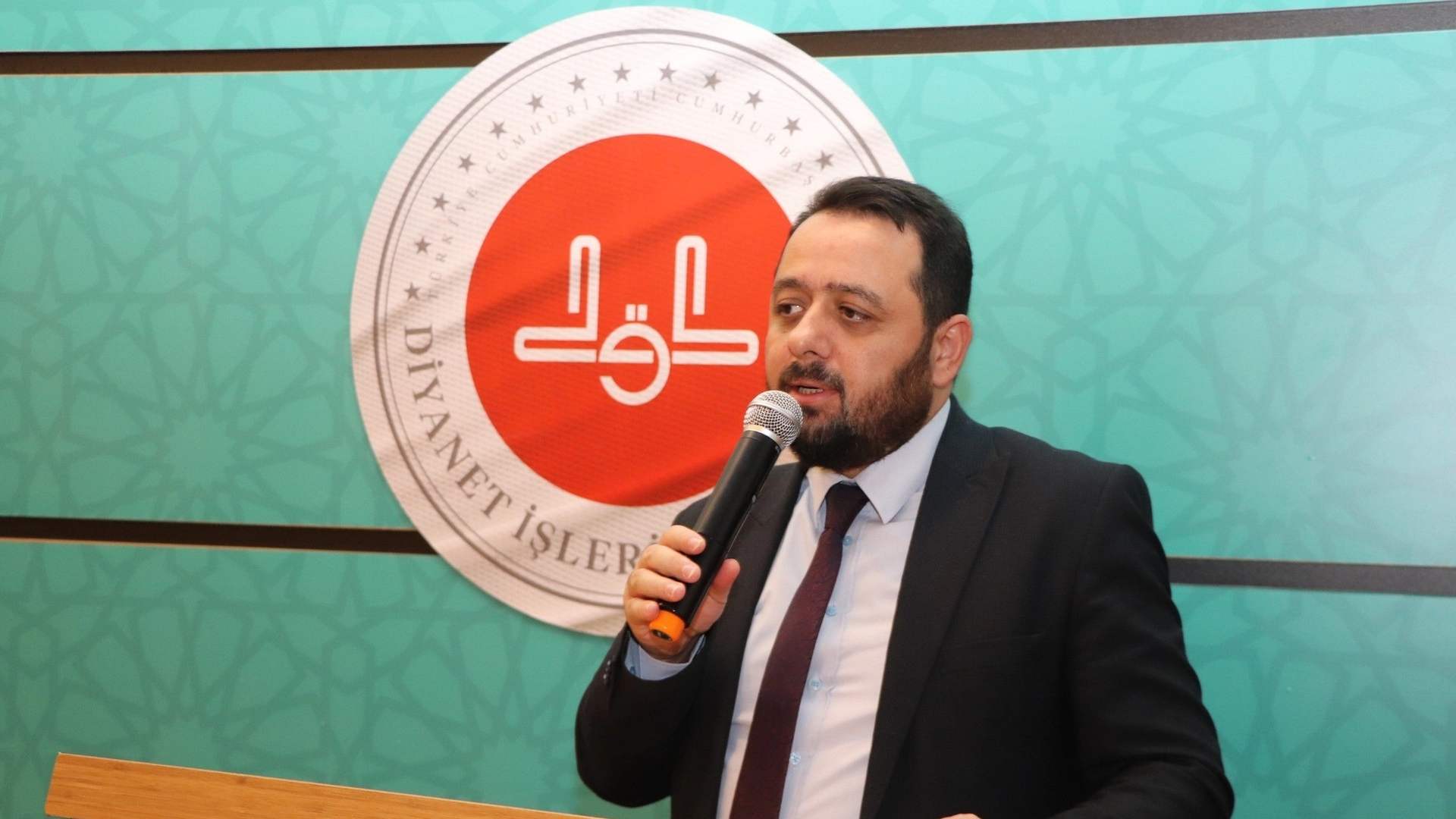 Kürsüde mikrofonla konuşan sakallı bir erkek, arkasındaki Diyanet İşleri Başkanlığı logolu panonun önünde duruyor.
