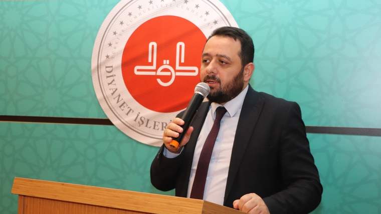 Kürsüde mikrofonla konuşan sakallı bir erkek, arkasındaki Diyanet İşleri Başkanlığı logolu panonun önünde duruyor.