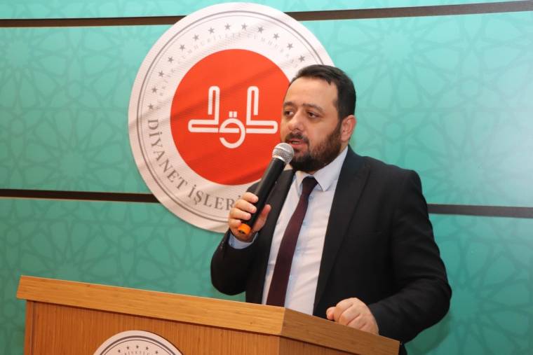 Kürsüde mikrofonla konuşan sakallı bir erkek, arkasındaki Diyanet İşleri Başkanlığı logolu panonun önünde duruyor.