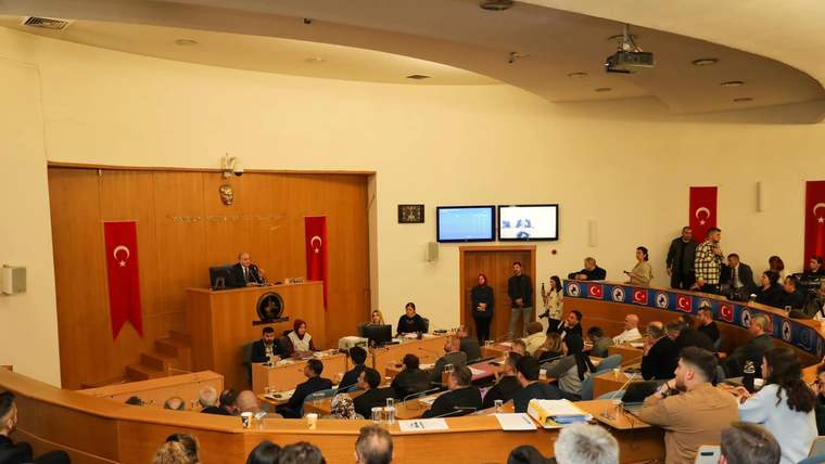 Türk bayrakları ve flamalarla süslenmiş salonda, kürsüde konuşan kişi ile oturan izleyiciler görülüyor.