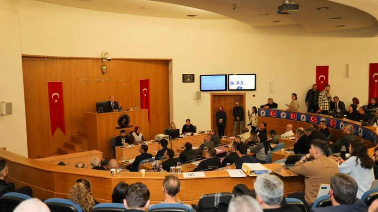 Türk bayrakları bulunan salonda, kürsüde konuşan kişi ve sıralarda oturan kalabalık görülüyor.