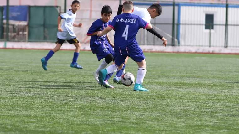 Yapay çim sahada dört çocuk futbolcu top için mücadele ederken, mor formalı oyuncunun sırtında 4 numara görülüyor.