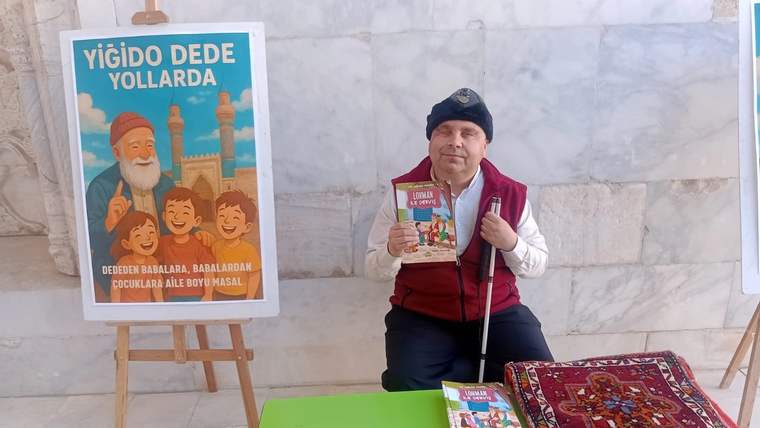 Bir kişi, elinde çocuk kitabı tutarak oturuyor; yanında masal kitabı afişi ve serili halı bulunuyor.