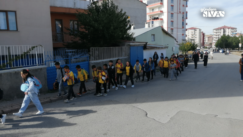 Sokakta yan yana yürüyen, bazıları sarı-siyah kıyafetler giyen çocuklar ve arkalarında yetişkinler görülüyor.