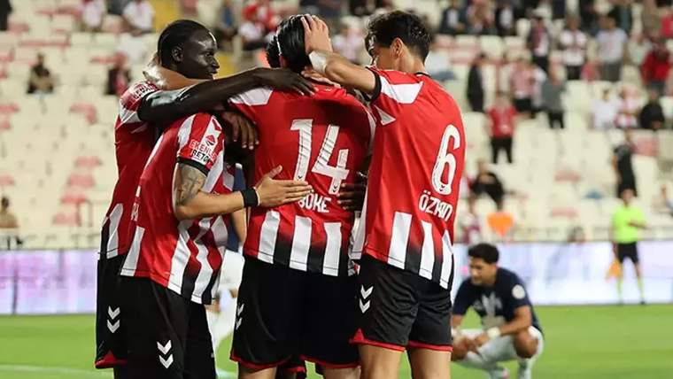 Kırmızı-beyaz formalar giyen dört futbolcu sahada birbirine sarılırken, arka planda bir oyuncu oturuyor.