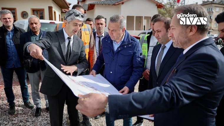 Bir grup erkek, bir harita üzerinde tartışıyor; arka planda inşaat alanı ve binalar var.