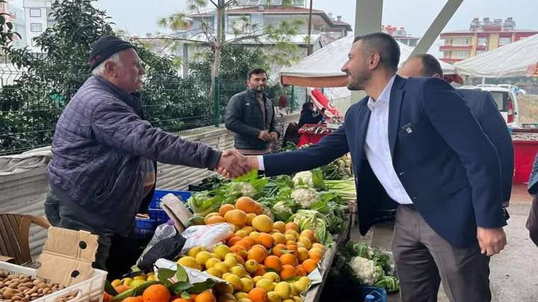 Pazar tezgâhının önünde iki erkek el sıkışıyor; tezgahta portakal, limon, marul ve karnabahar görülüyor.