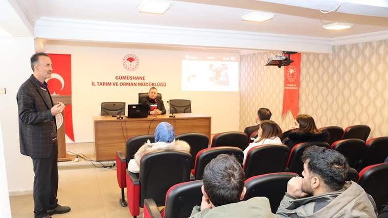Toplantı salonunda kürsüde konuşan kişi, oturan katılımcılar ve arkada sunum ekranı görülüyor.