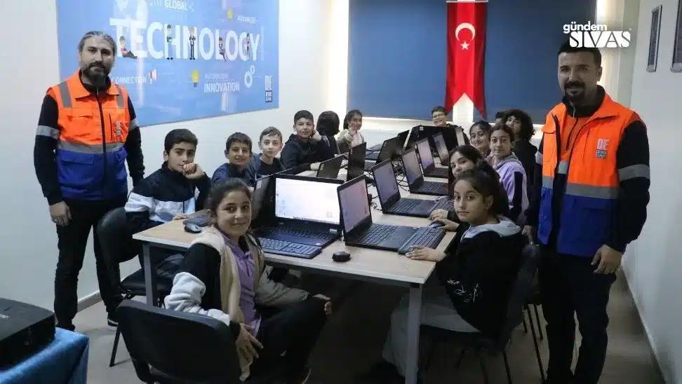 Bir grup öğrenci ve öğretmen, bilgisayarların bulunduğu bir sınıfta gülümseyerek poz veriyor.