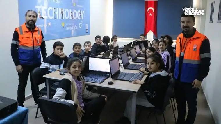 Bir grup öğrenci ve öğretmen, bilgisayarların bulunduğu bir sınıfta gülümseyerek poz veriyor.