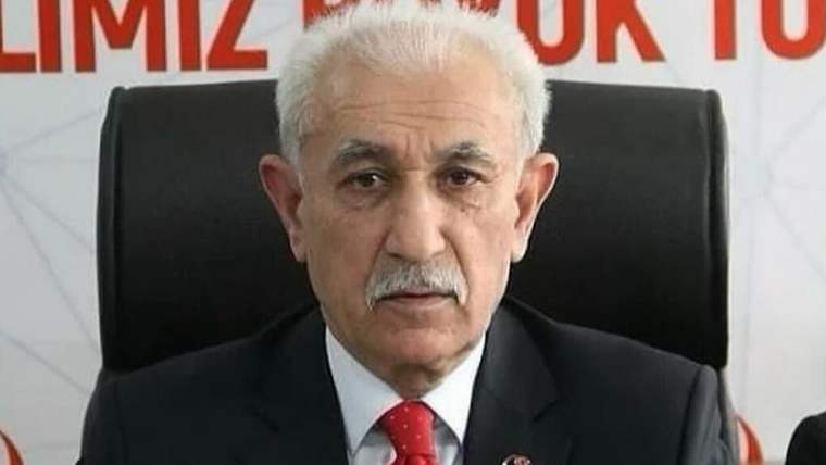 Beyaz saçlı ve bıyıklı bir kişi, siyah takım elbise ve kırmızı kravatla siyah koltukta oturuyor.