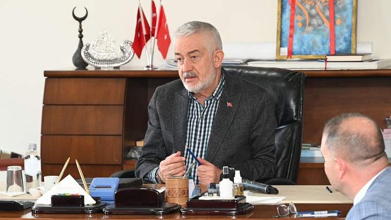 Masada oturan bir kişi elinde kalem tutuyor; karşısında başka bir kişi ve arka planda Türk bayrakları bulunuyor.