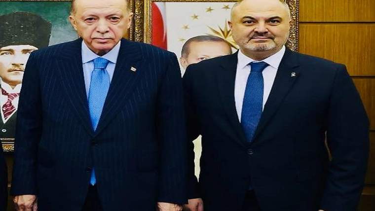 Takım elbiseli iki erkek, arkalarında çerçeveli portreler bulunan ahşap kaplı bir odada yan yana duruyor.