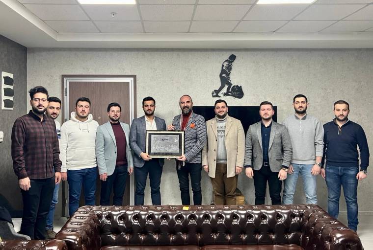 Bir odada dokuz erkek ayakta duruyor; ortadaki iki kişi çerçeveli bir plaketi birlikte tutuyor.