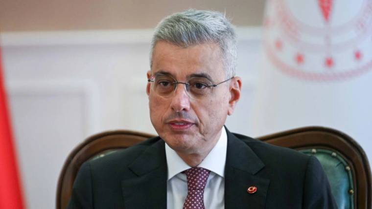 Gözlüklü, takım elbiseli ve kravatlı bir kişi, arkasında amblem bulunan koltukta oturuyor.