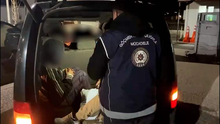 Gece bir araçta oturan kişiler ve arka kapı önünde, üzerinde "Göçmen Kaçakçılığıyla Mücadele" yazan görevli görülüyor.