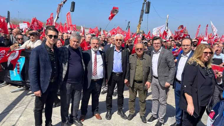 Kırmızı bayraklar taşıyan kalabalığın önünde, yan yana duran bir grup kişi kameraya bakıyor.