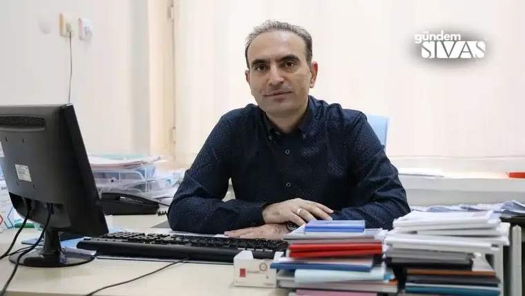 Masa başında oturan bir kişi, bilgisayar ve üst üste dizilmiş dosyaların önünde kameraya bakıyor.