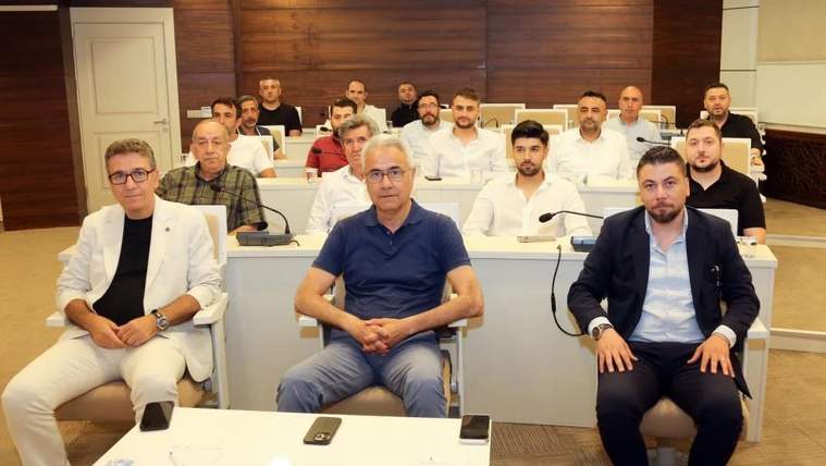 Bir toplantı salonunda oturan sigorta acenteleri, masanın etrafında sıralanmış. Arka planda tahta kaplama duvar var.