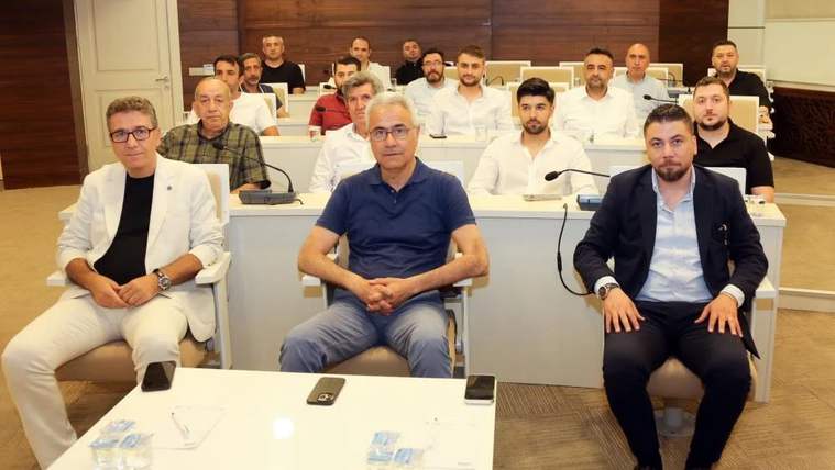 Bir toplantı salonunda oturan sigorta acenteleri, masanın etrafında sıralanmış. Arka planda tahta kaplama duvar var.