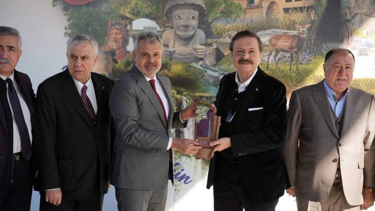 Beş erkek, duvar resminin önünde duruyor; ortadaki iki kişi ellerinde plaket tutuyor.