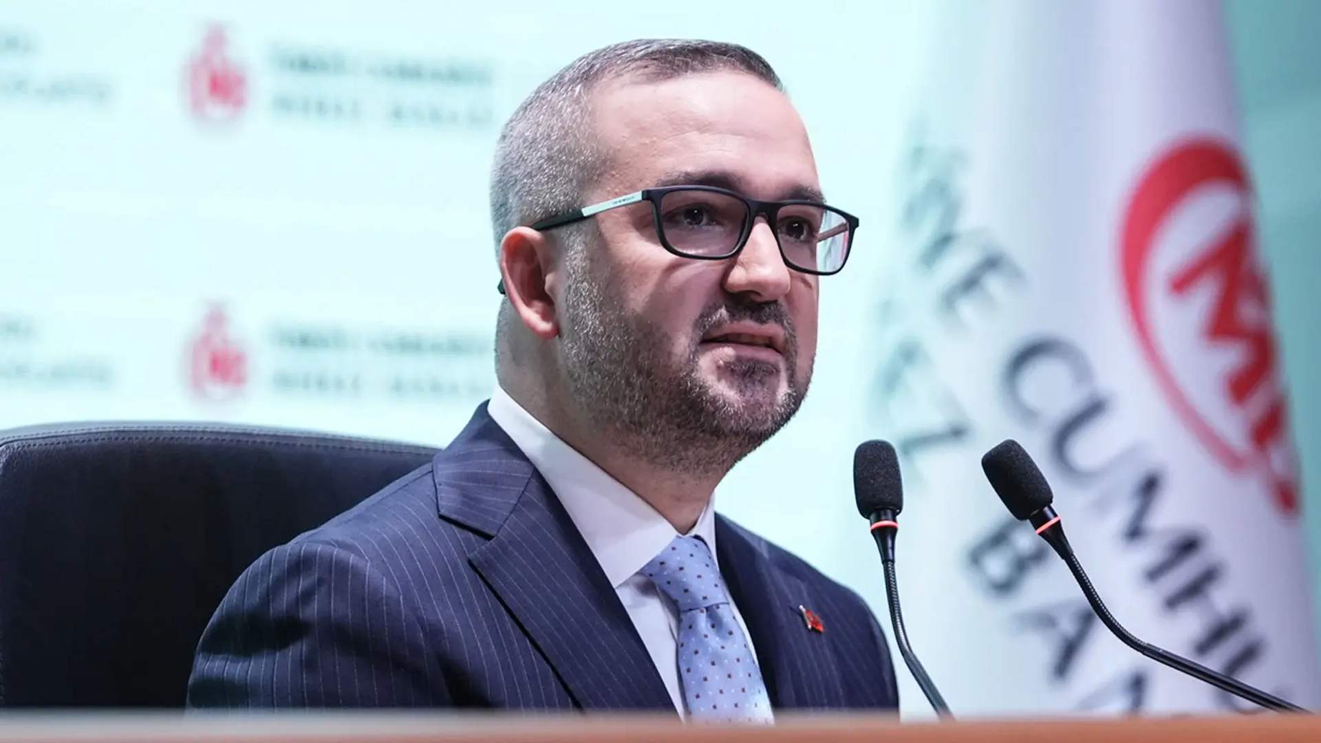 Gözlüklü bir kişi, takım elbiseyle iki mikrofonun önünde konuşurken; arkada yazılı bir pano bulunuyor.
