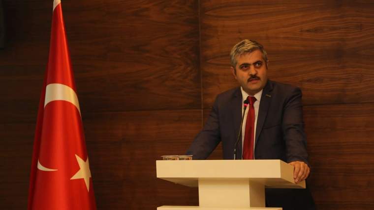 Kürsüde konuşan bir kişi, yanında Türk bayrağı bulunan ahşap panelli salonda görülüyor.