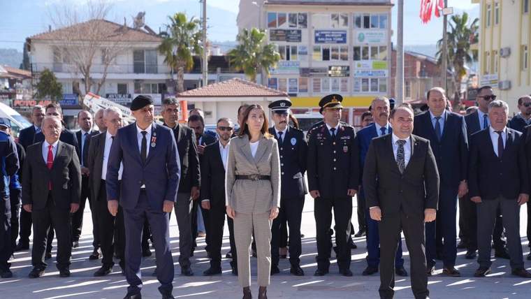 Meydanda takım elbiseli kişiler ve üniformalı görevliler yan yana yürürken, arkada binalar ve Türk bayrakları görülüyor.