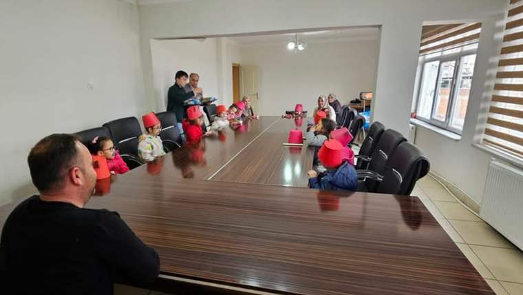 Toplantı masası çevresinde oturan çocuklar ve yetişkinler, iç mekânda karşılıklı duruyor.