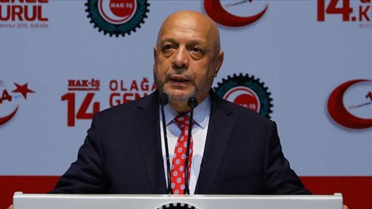 Takım elbiseli bir kişi, mikrofon önünde konuşurken arkasında sendika logoları ve yazıları görülüyor.