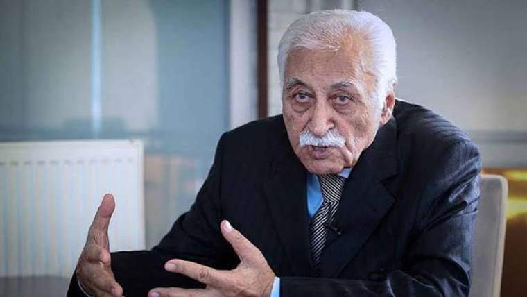 Koyu renkli takım elbise giymiş, beyaz saçlı ve bıyıklı bir kişi, el hareketleriyle konuşuyor.
