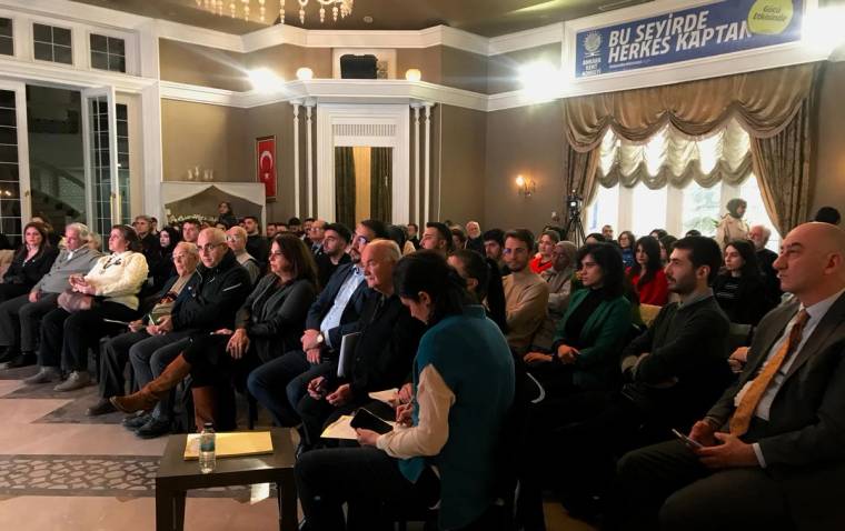 Süslü bir salonda, Türk bayrağı ve duvar afişi önünde oturan kalabalık izleyici grubu.