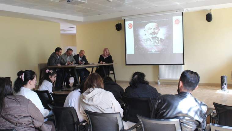 Projeksiyon ekranı önünde masada oturan dört kişi ve onları izleyen öğrencilerden oluşan grup görülüyor.