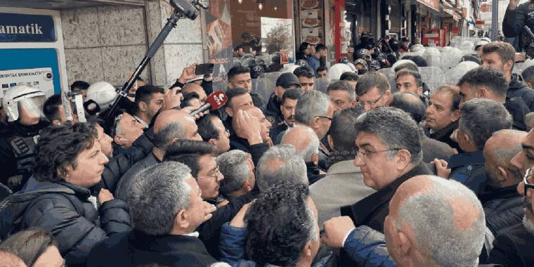Sokakta toplanan kalabalık arasında mikrofon ve kameralarla konuşan gözlüklü bir kişi görülüyor.