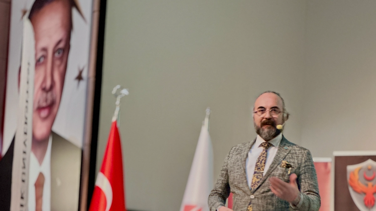 Sahnede mikrofonlu bir kişi el hareketiyle konuşurken yanında Türk bayrağı ve beyaz logolu bayrak görülüyor.