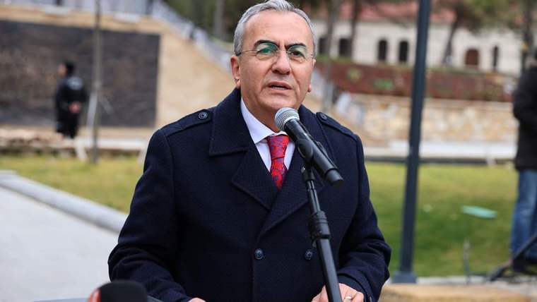 Mikrofonun önünde konuşan bir kişi, arka planda park alanı görünmekte.