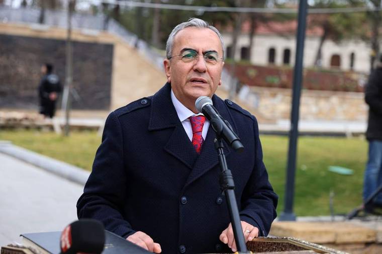 Mikrofonun önünde konuşan bir kişi, arka planda park alanı görünmekte.